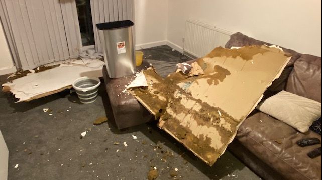 A large piece of collapsed ceiling, which has fallen onto a brown leather sofa and has left shards of debris across the grey carpet of the living room. There is a silver bin in the background surrounded by what looks like another piece of fallen ceiling and a collapsible bucket. 