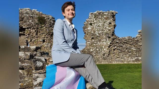 Gwion Williams smiling at the camera in the ruins of a castle with the blue, pink and white trans flag laid out next to him