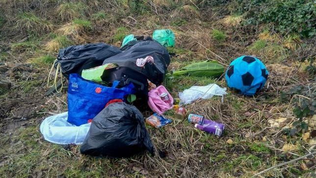 A pile of discarded shopping bags, black refuse bags and toys such as a football, discarded in a field near a wooden fence