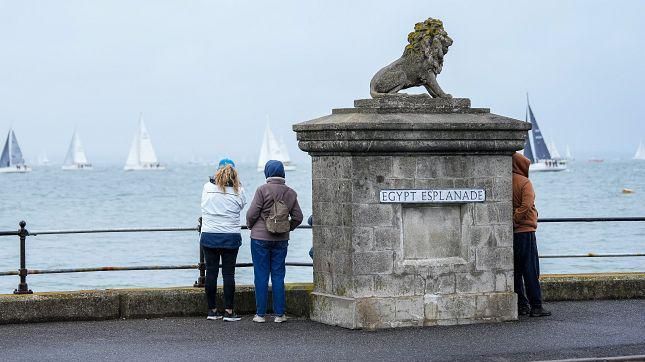 People in Cowes on the Isle of Wight stood by a signpost for Egypt Esplanade looking out at the sea where yachts with blue and white sails can be seen in the distance