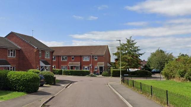 Bibury Road in Gloucester with houses made of red brick with slate roofs, alongside a road. There are also areas of lawn and trees visible