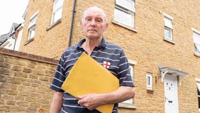 David Riley standing in front of his former home, wearing blue top with pink stripes and holding orange folder. 