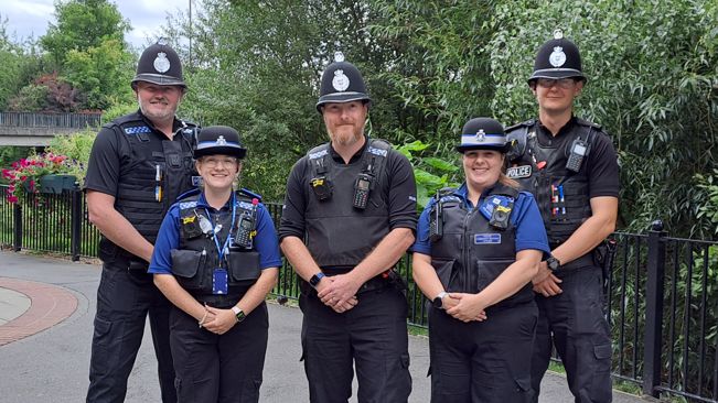 A group of five police officers, three men and two women, stand together in a pedestrianised area smiling at the camera.