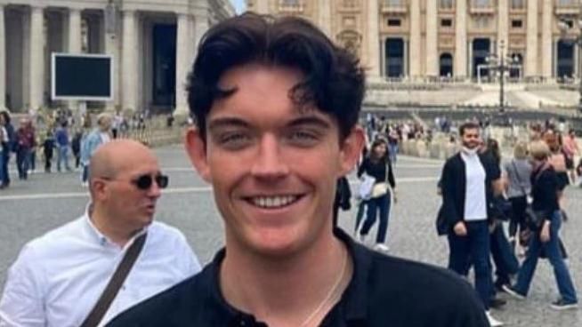 A smiling Charlie Pearson in what appears to be a square with people mingling in the background. He has dark brown hair and is wearing a dark top