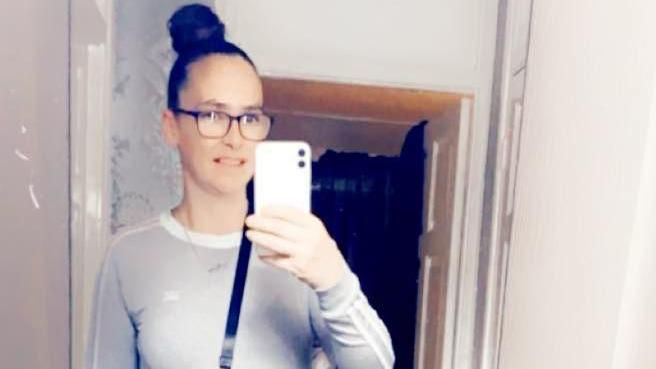 Kelly wearing a grey top and black glasses with her hair in a bun. She is holding a white phone as she smiles in a mirror.