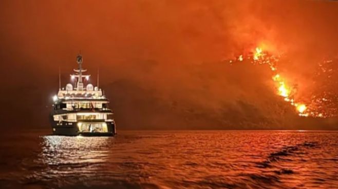 A photo of a luxury yatch on the water, with a raging fire on the land in the distance.