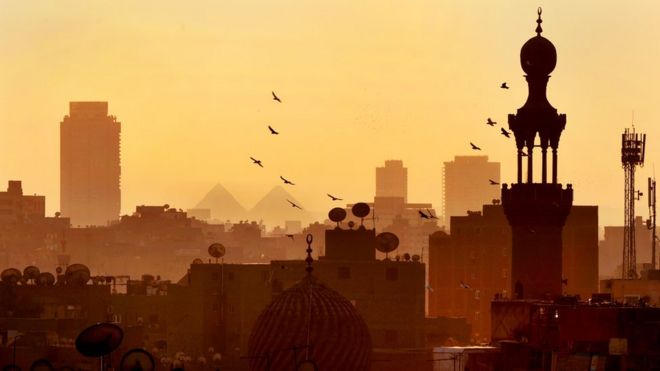 Prédios e construções antigas ao entardecer no Cairo, de onde se vê ao fundo duas pirâmides
