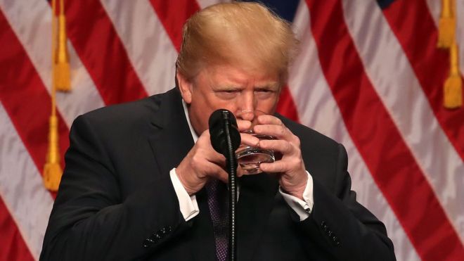 Президент США Дональд Трамп пьет из стакана воды, который он держит двумя руками, во время выступления в здании Рональда Рейгана 18 декабря 2017 года в Вашингтоне, округ Колумбия.