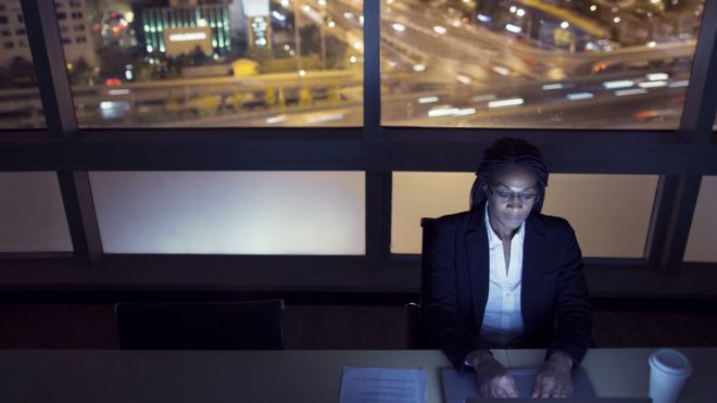 Mulher digita em computador dentro de escritório, com vista da cidade grande à noite na janela