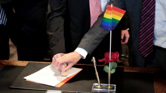 Однополая пара ставит на стол документы о браке с радужным флагом