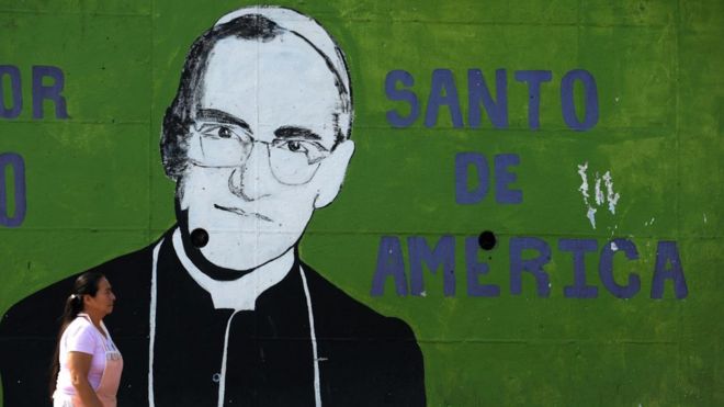 Imagem mostra mulher passando em frente à parede pintada com imagem do monsenhor Oscar Arnulfo Romero, o novo santo da Igreja