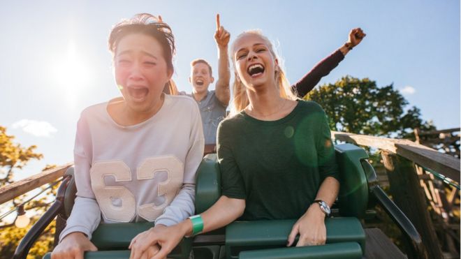 Two women scream as they ride a rollercoaster, with friends in background
