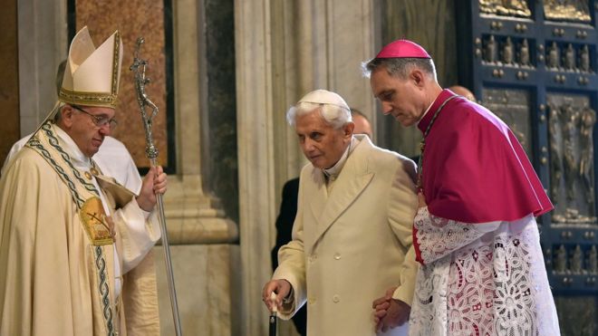 Почетному Папе Бенедикту XVI помогает префект папского двора Георг Гаенсвайн (справа) пройти через Святую Дверь, пока папа Франциск (слева) смотрит на