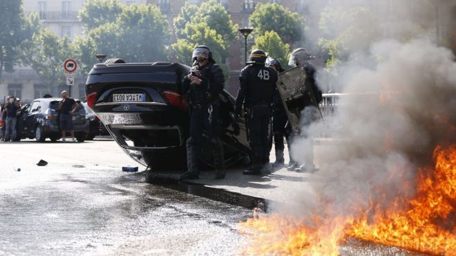 Дым поднимается от пожара, который горит рядом с французским спецназом CRS, стоящим возле перевернутой машины, так как таксисты блокируют Порт Майо в Париже 25 июня 2015 года.