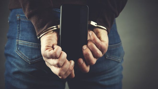 A handcuffed man holds a mobile phone behind his back
