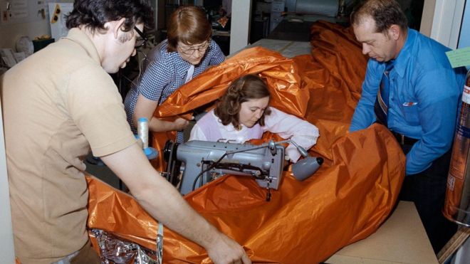 Em foto antiga, já colorida, Aylene Baker aparece sentada diante de uma máquina de costura, cercada por material (tipo tecido) volumoso segurado com a ajuda de várias pessoas
