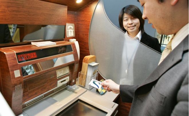 Человек стоит перед японским банкоматом, держа в руках банковскую карту Shinsei, а сотрудник банка смотрит на