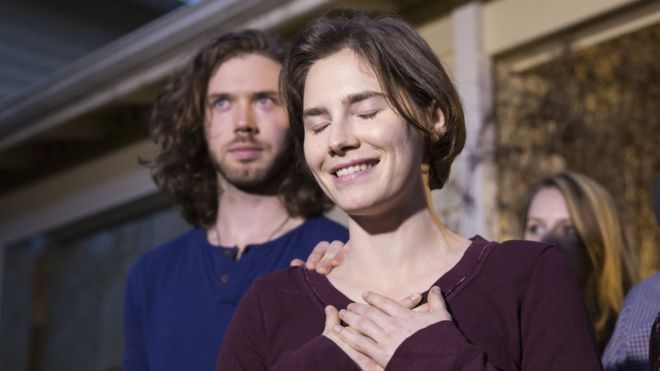 Amanda Knox, eyes closed, smiles and clasps her hands over her heart, while her fiance, a bearded and long-haired young man, places a hand on her shoulder