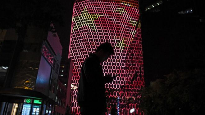 Мужчина смотрит в свой телефон рядом с гигантским изображением китайского национального флага на стене здания в Пекине в 2017 году