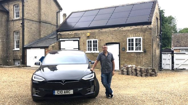 Адам Кортни, стоящий рядом с автомобилем Тесла перед домом, установит солнечные батареи