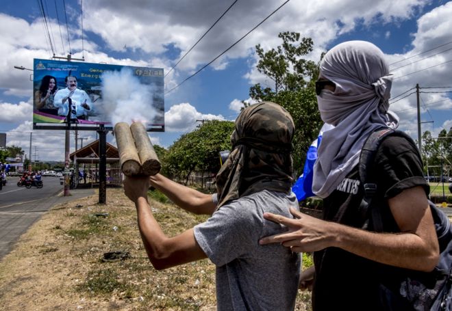 Студенты в масках стреляют своими самодельными минометами по большому плакату президента Даниэля Ортеги и его жены, вице-президента Росарио Мурильо