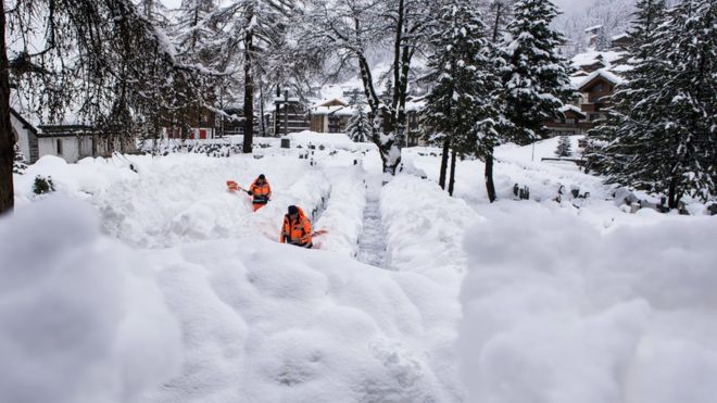 Двое рабочих в флуоресцентном оранжевом цвете убирают снег с дорожки в Церматте - он легко до пояса местами, когда они сгребают его в сторону