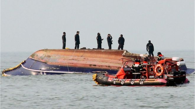 Six members of the South Koran coastguard stand on top of an overturned hull of the fishing boat, in the middle of the water, while their own rubber dinghy waits nearby