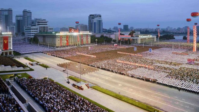 В Пхеньяне на переполненном квадрате изображены люди, одетые в одинаковые цвета в аккуратных прямоугольных единицах на тысячу человек, собравшиеся перед зданиями, украшенными политическими знаменами