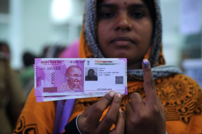 Индийская женщина позирует с новыми банкнотами 2000 рупий, ее удостоверением личности Аадхаара и пальцем, нанесенным несмываемыми чернилами, после обмена снятыми 500 и 1000 рупий банкнотами в банке в Ченнае 17 ноября 2016 года