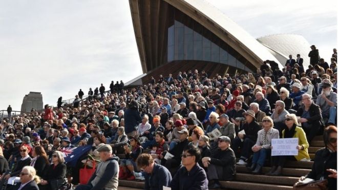 Люди сидят на ступеньках Сиднейского оперного театра, чтобы посмотреть поминальную службу