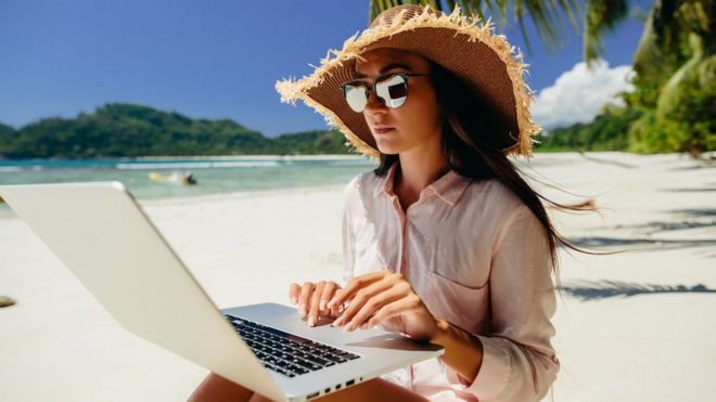 Una mujer con lentes de sol y sombrero trabaja con una computadora en una playa.