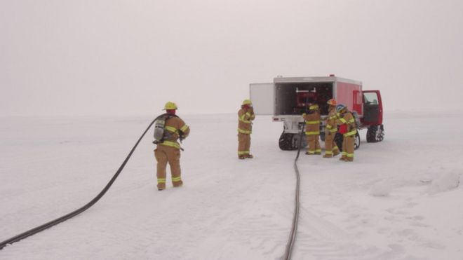 Пять пожарных работают из машины, припаркованной на льду. За ними тянутся шланги, а небо белое.