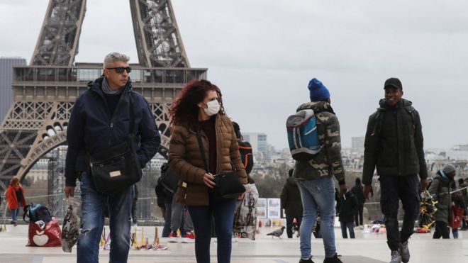 Pessoas em frente à torre eiffel, uma delas usa máscara