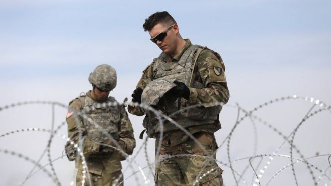 Dois soldados fardados olham para seus equipamentos com ar compenetrado, atrás de uma cerca de arame farpado