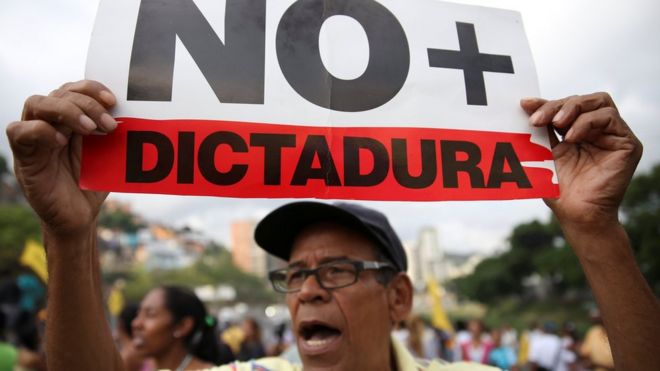 Сторонник оппозиции держит плакат с надписью «Нет больше диктатуры». выкрикивает лозунги, когда он блокирует шоссе во время протеста против правительства президента Венесуэлы Николаса Мадуро в Каракасе