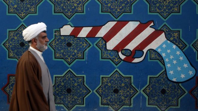 An Iranian clerric walks past a mural on the wall of the former US embassy