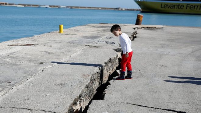Child peers at damaged pier at port on Zakynthos