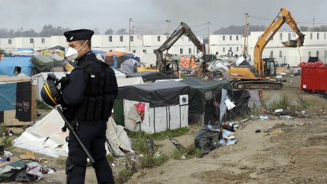 Офицер полиции стоит на страже, в то время как 26 октября 2016 года механические землекопы начинают разрушать укрытия во временном лагере мигрантов, известном как Джунгли, недалеко от Кале, на севере Франции. Было неясно, кто зажег десятки пожаров, которые вспыхнули в одночасье и до утра