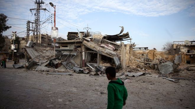 Criança caminha perto de construções danificadas por ataques na cidade de Douma