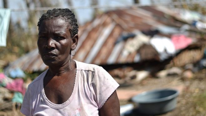 Una mujer haitiana con su casa en ruinas al fondo.