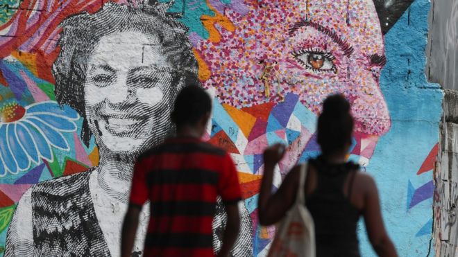 Sombras de dois pedestres passando em frente a painel com homenagens a Marielle Franco, retratada em grafites