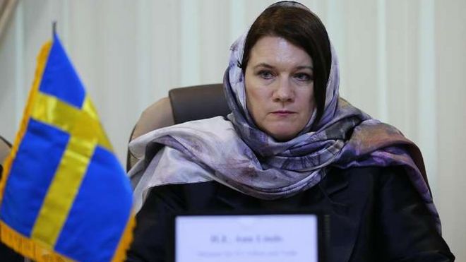 Ann Linde wearing a light purple headscarf, sitting at a desk with a small Swedish flag in Tehran, Iran