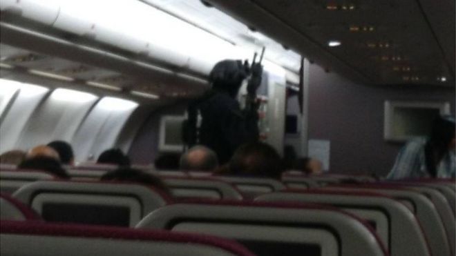 На расплывчатой ??фотографии изображены пассажиры, сидящие в самолете, в то время как тяжело вооруженный человек в полном тактическом снаряжении и что-то похожее на винтовку идет к передней части самолета