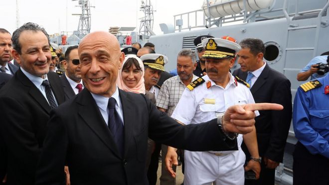 Марко Миннити смеется вместе с министром обороны Ливии (поддерживаемого ООН правительства) в гавани рядом с некоторыми ливийскими судами береговой охраны, в то время как военно-морской персонал также можно увидеть поблизости
