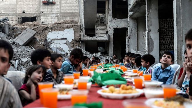 Дети сидят за столом, ожидая ифтара, вечернего обеда в конце ежедневного поста Рамадан на закате, рядом с обломками и разрушенными домами, которые были повреждены в результате воздушных ударов, в Дума, Сирия, 17 июня 2017 года.