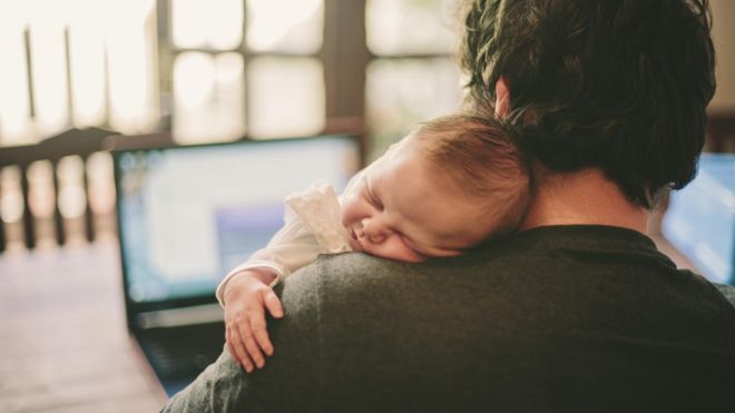 Father working on his laptop while his newborn baby girl sleeps on his shoulder