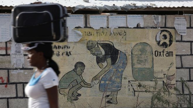 A woman walks carrying a suitcase on her head next to an Oxfam sign on the outskirts of Port-au-Prince, Haiti