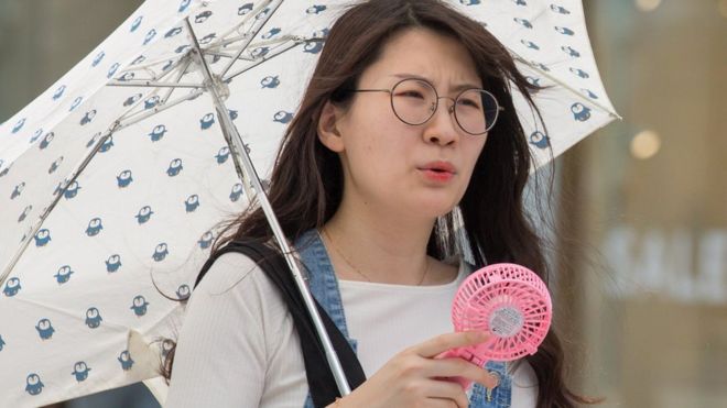 A woman in Japan holds a pink fan to herself, carrying an umbrella to avoid heat