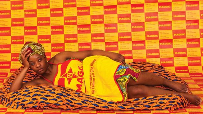 A woman lies on a soft blanket, looking into the camera, wearing Maggi stock cube branded dress, behind her a wallpaper made of the Maggi logo