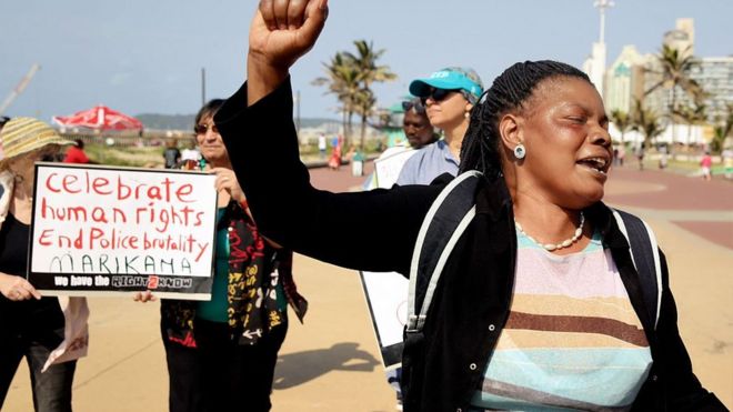 Протестующий поднимает кулак, когда другие держат плакат с надписью «Отпразднуйте права человека, покончите с жестокостью полиции» во время празднования третьей годовщины кровавой расправы на Марикане на северном пляже в Дурбане 16 августа 2015 года.